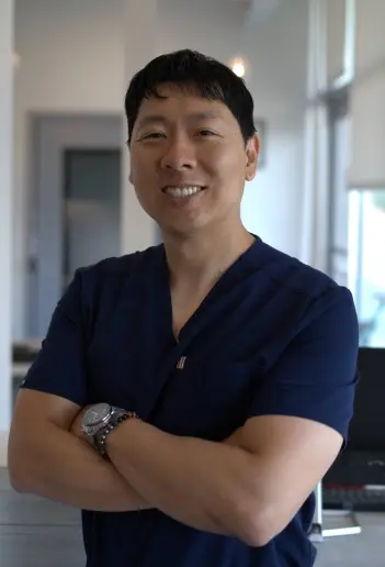 Smiling dentist in navy scrubs with arms crossed in modern dental office.